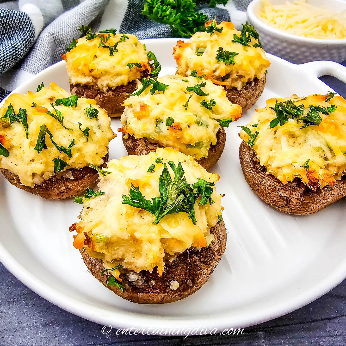 Five baked stuffed mushrooms topped with melted cheese and fresh parsley are arranged on a white plate, with a small bowl of grated cheese in the background.