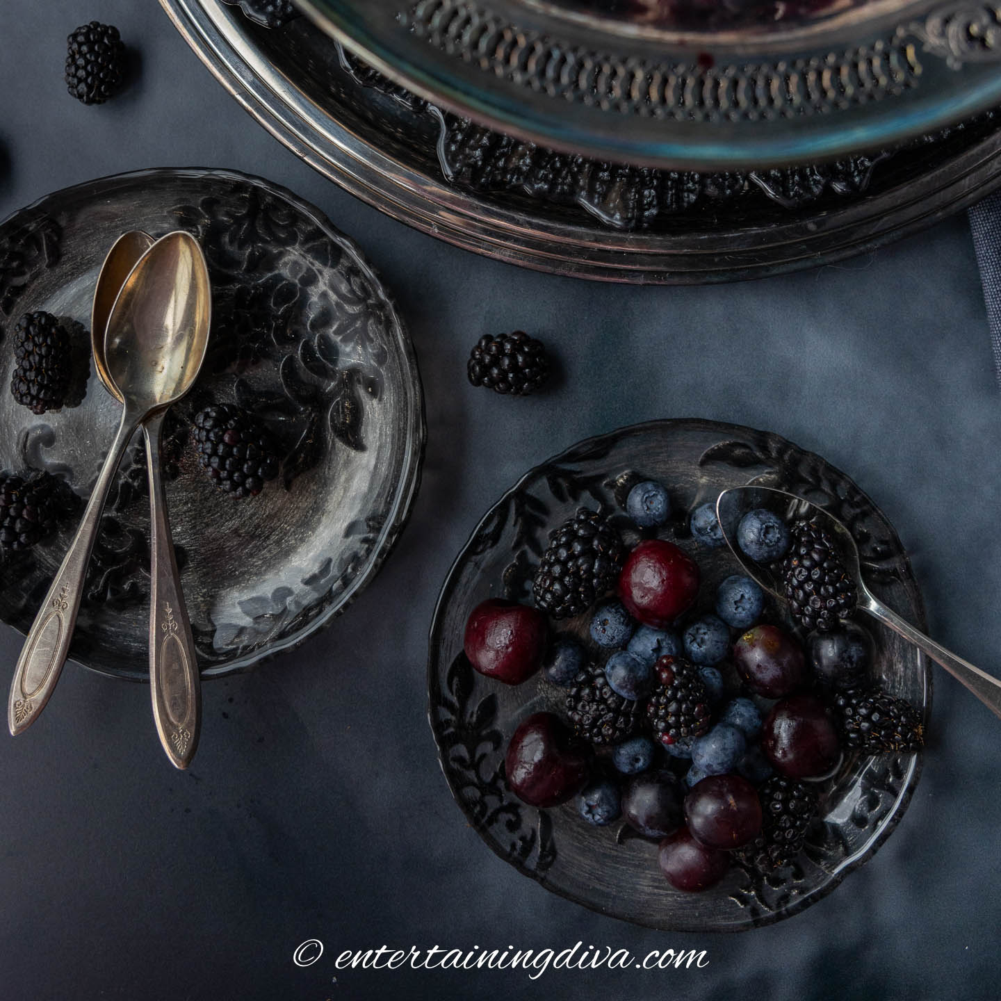 dark Halloween fruit salad on a grey and black plate beside a stack of plates with silver spoons