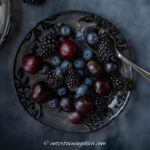 A dark glass plate with blackberries, blueberries, cherries, and grapes, with a small spoon on the side. The plate rests on a dark surface.