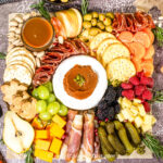 Charcuterie board with crackers, brie, cured meats, cheese cubes, pickles, fruit slices, berries, nuts, olives, and a bowl of dip, arranged neatly on a textured surface.