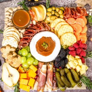 Charcuterie board with crackers, brie, cured meats, cheese cubes, pickles, fruit slices, berries, nuts, olives, and a bowl of dip, arranged neatly on a textured surface.