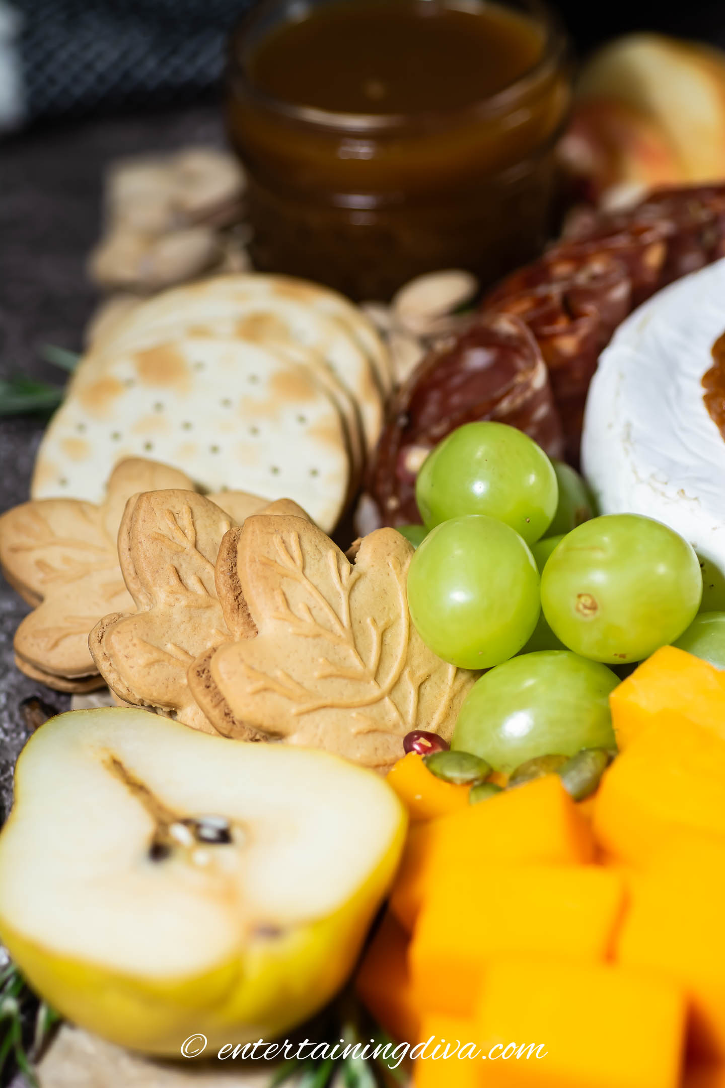 Close up of a pear, cheddar cheese cubes, green grapes, maple leaf cookies, crackers and salami on a fall charcuterie board