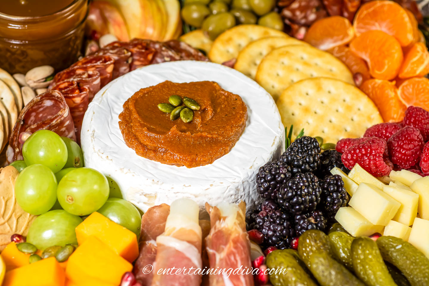 close up of the center of a fall charcuterie board with brie, pumpkin butter, grapes, berries, crackers, cheddar cheese, prosciutto wrapped mozzarella, salami and pickles