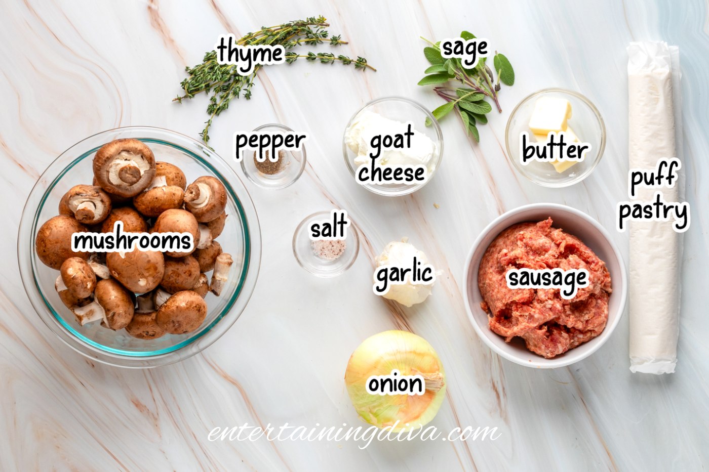 Ingredients for a mushroom sausage puff pastry bites recipe arranged on a counter, including mushrooms, thyme, sage, goat cheese, butter, puff pastry, sausage, onion, garlic, salt, and pepper.