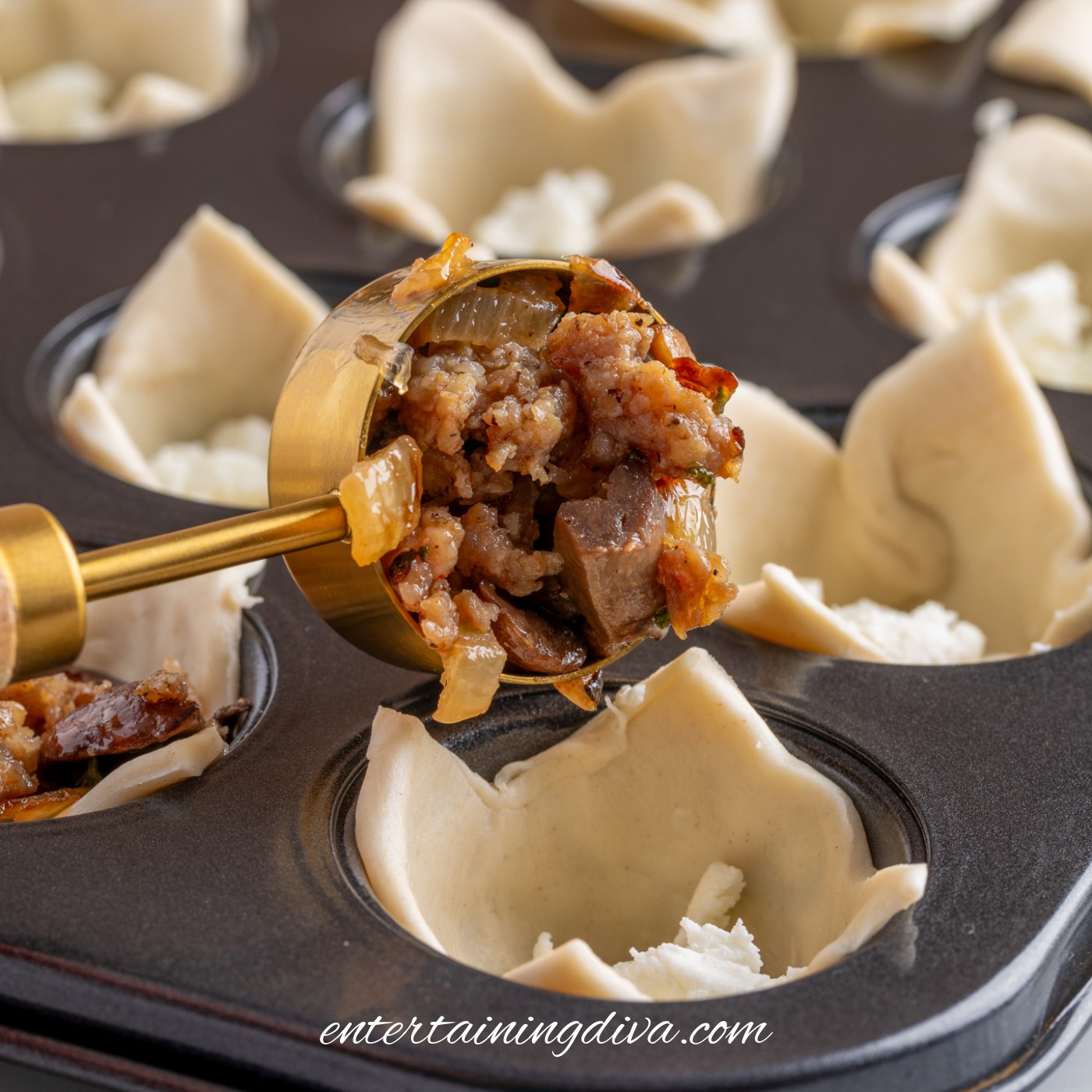 A metal scoop adds cooked sausage filling to pastry cups in a muffin tin; bits of white cheese are visible in the cups.