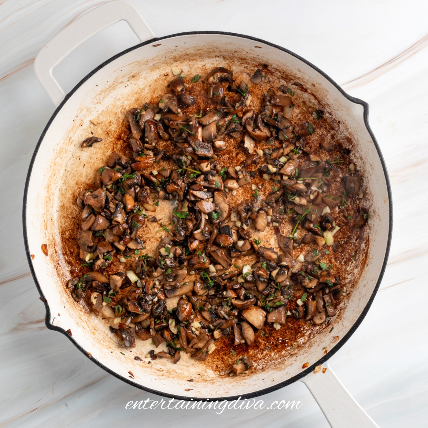 Chopped mushrooms, herbs, onions and garlic sautéed in a large white pan with browned bits on the bottom.