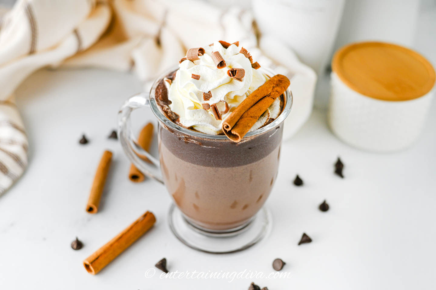 rumchata hot chocolate in a glass mug with whipped cream, chocolate shavings, chocolate chips and cinnamon sticks in the background