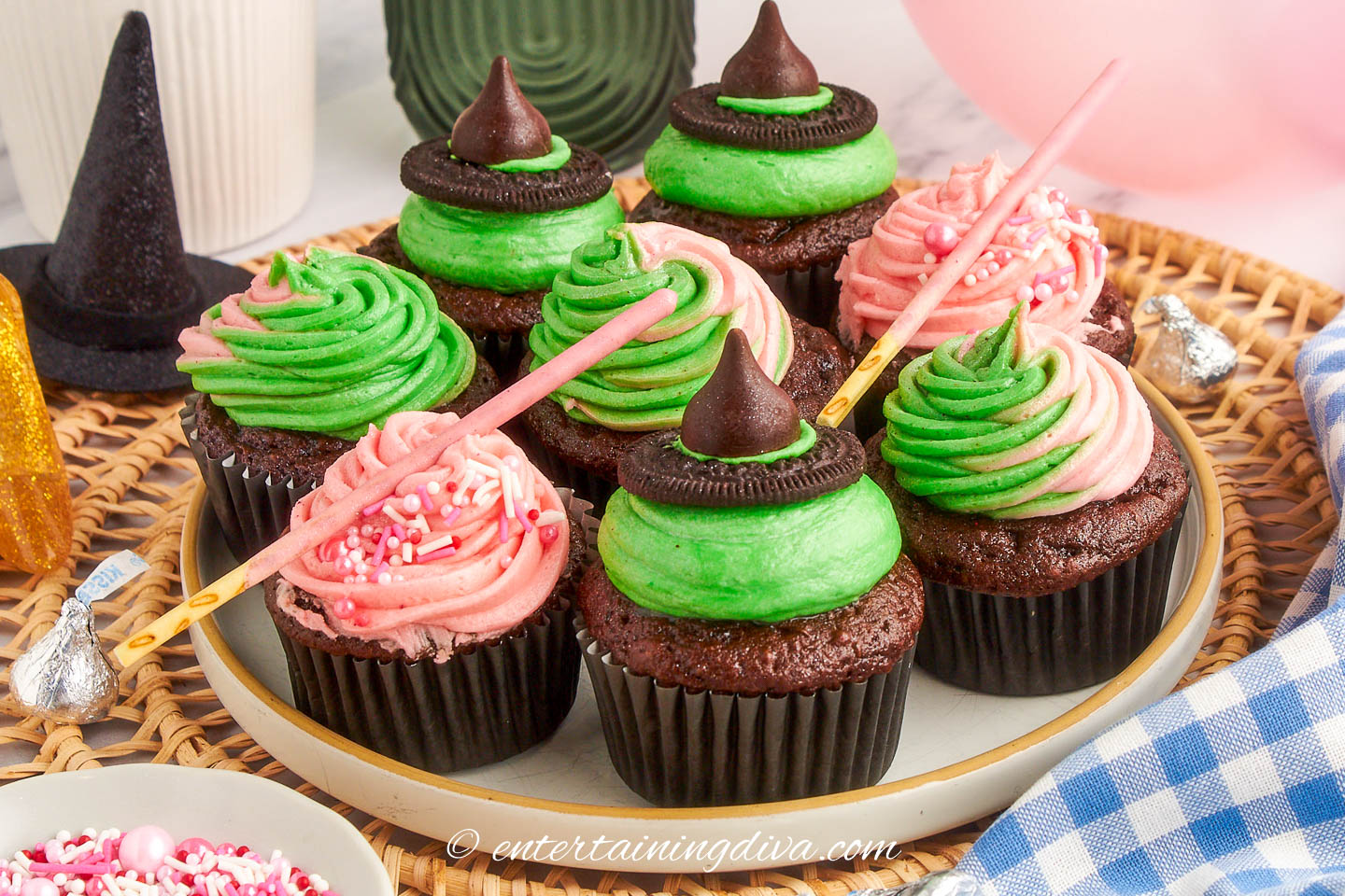 A mixture of 3 different Wicked cupcakes designs on a plate - green ones with a witch hat for Elphaba, pink ones with a wanda for Glinda and some topped with pink and green swirled frosting