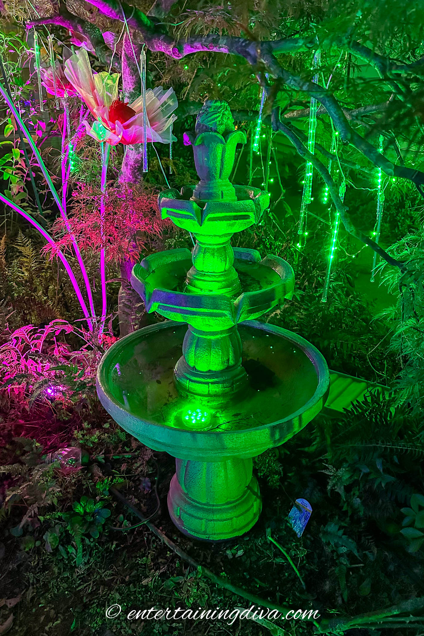 A 3-tier water fountain lit with green flood lights and green meteor lights in a Halloween Emerald City yard display