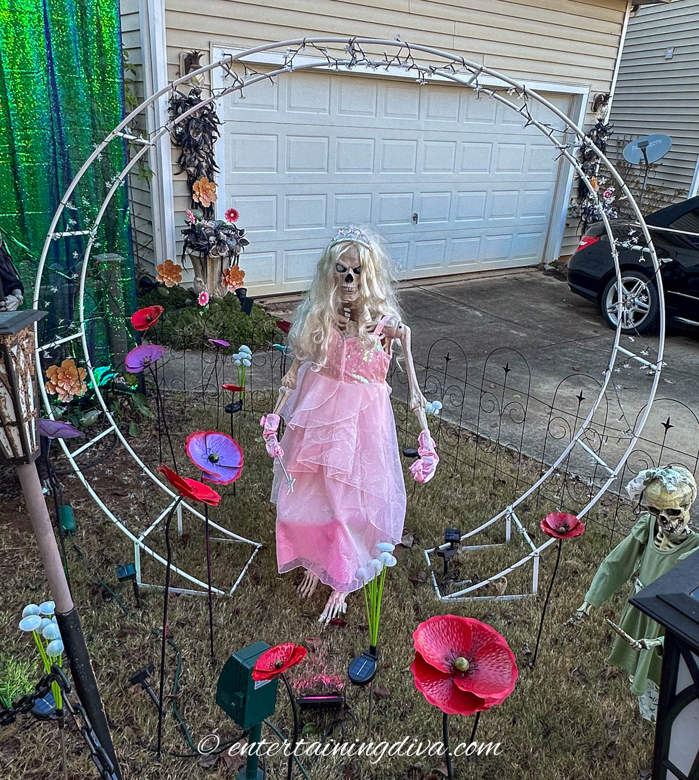 A skeleton dressed in a pink gown stands under a metal arch in a yard decorated with artificial flowers and Halloween props.