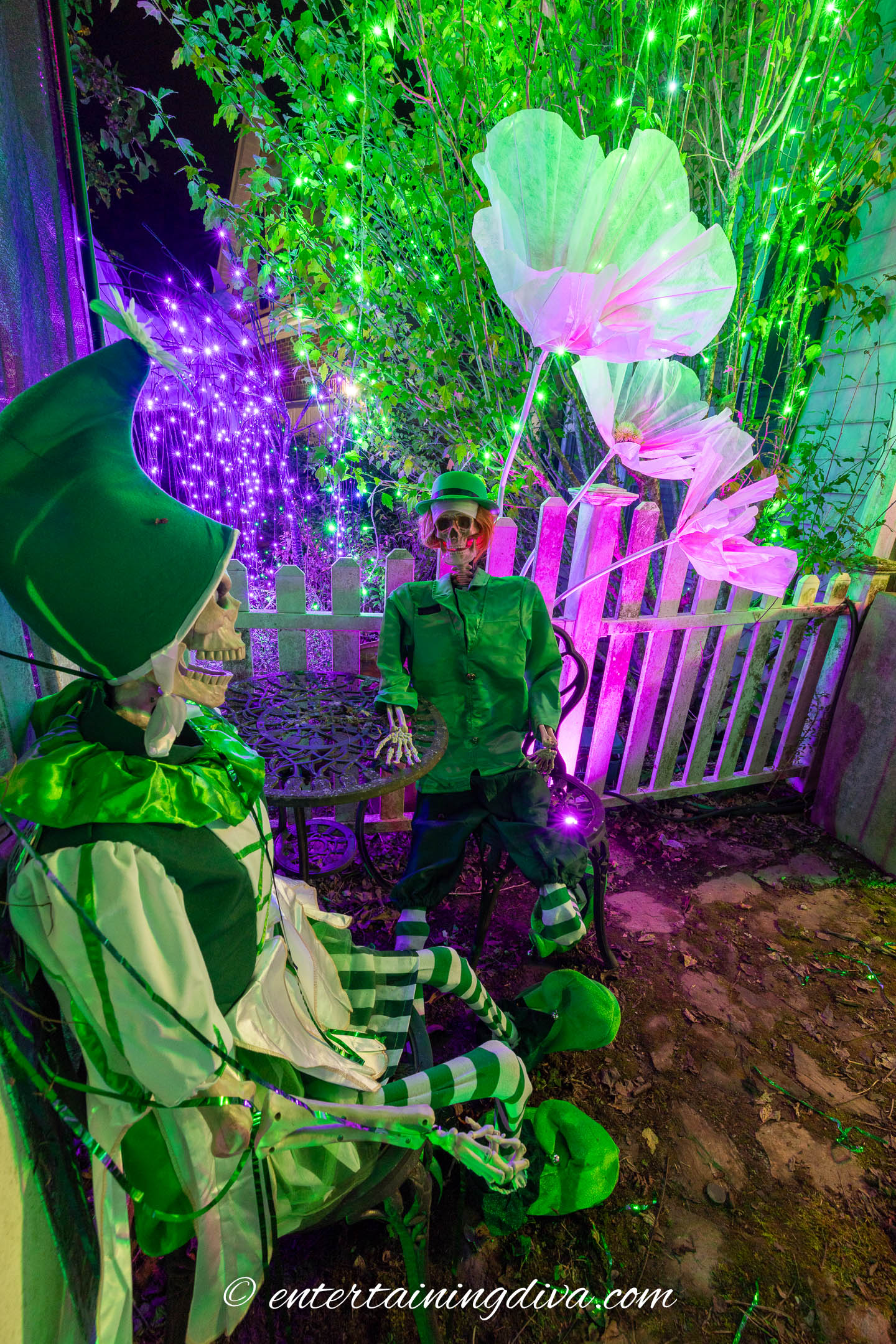 Skeletons dressed as Emerald City people in front of large artificial pink flowers in a Wicked-themed Halloween yard display at night