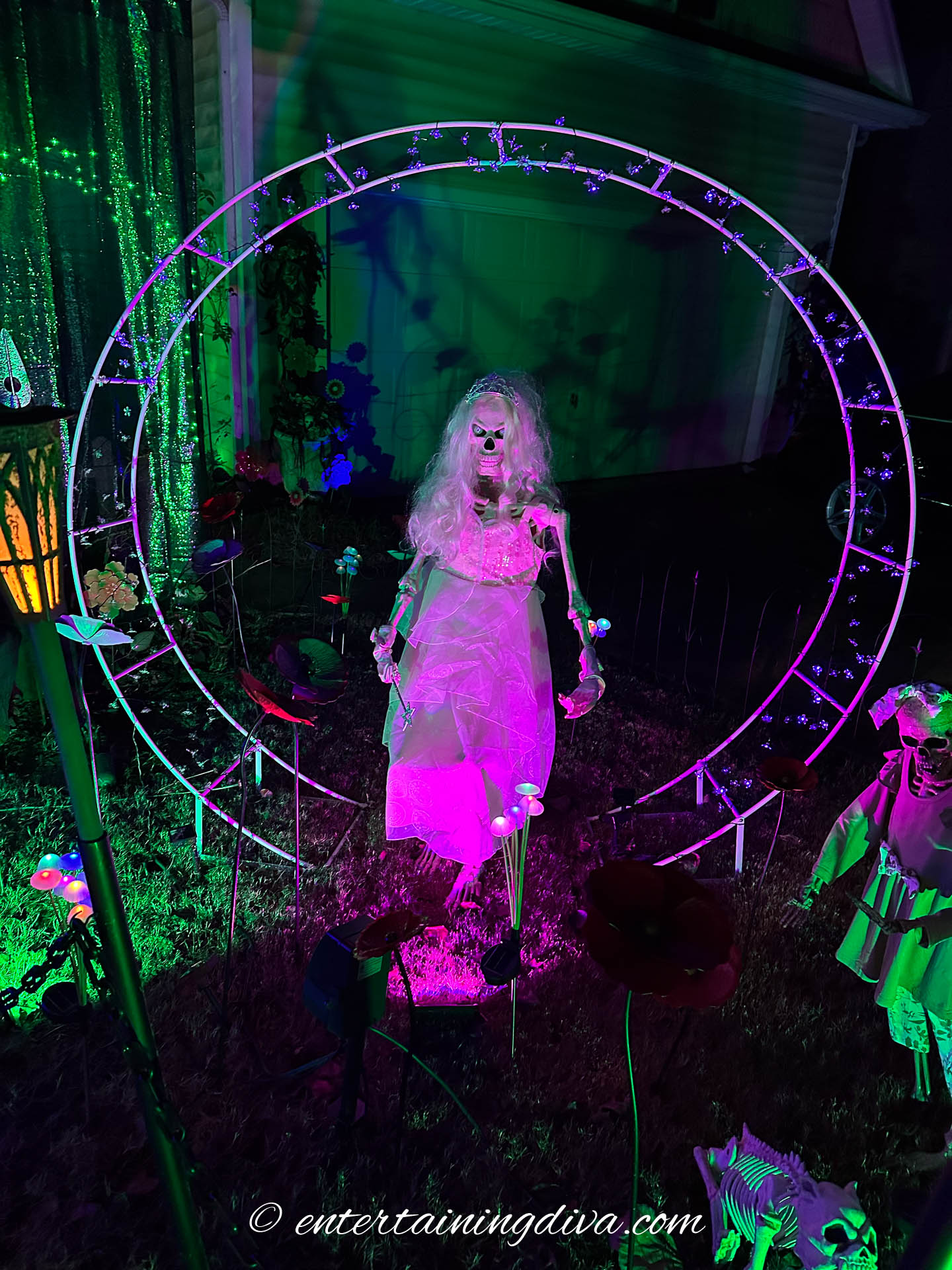 A skeleton dressed like Glinda in a pink arch 'bubble" in a Wicked Halloween yard display at night