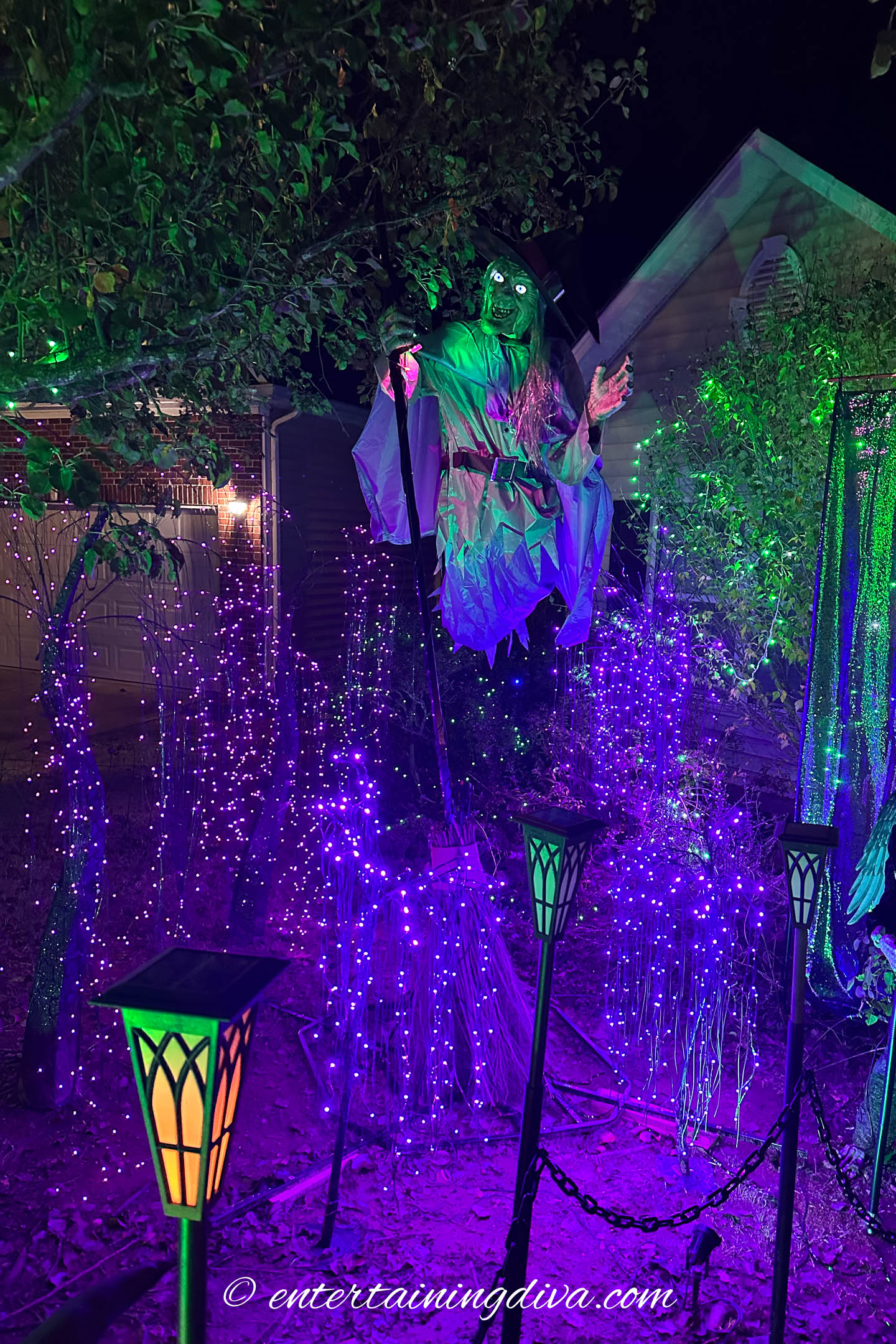 A flying witch prop surround by purple spooky trees in a Halloween front yard at night