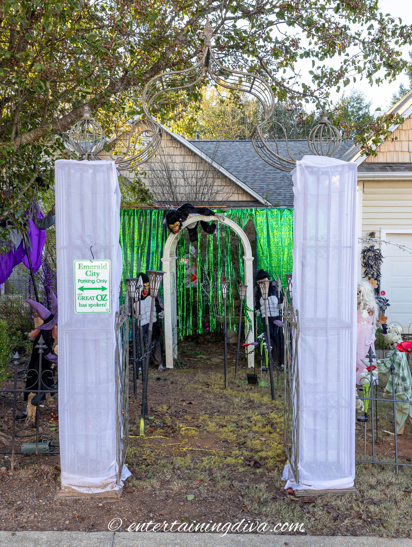 Outdoor Halloween display with green tinsel curtain, witch figure on archway, “Emerald City” sign, two white pillars, and spooky decorations in a front yard.