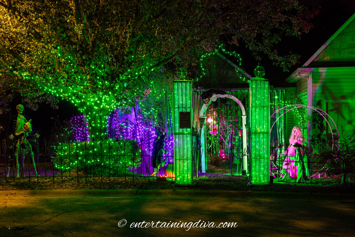A house decorated for Halloween with green and purple lights, skeleton figures, and Halloween props in the yard at night.