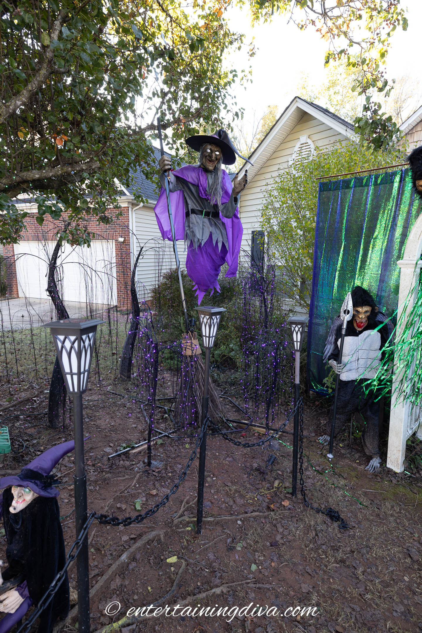 Three witch decorations are displayed in a yard with a light post, purple lights, and a wire fence. One witch is elevated on a broom, and two others sit near the ground, surrounded by trees.