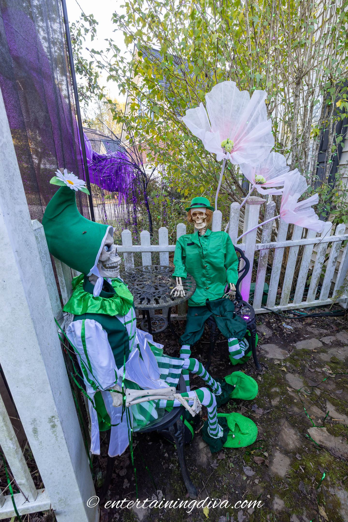 Two skeletons dressed in green costumes and hats sit on chairs in a garden area, holding oversized white flowers near a white picket fence.