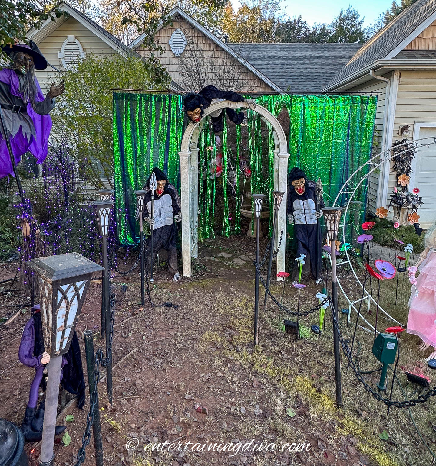 Front yard Halloween display with witches, skeletons, green tinsel backdrop, and themed decorations near a house and garden archway.