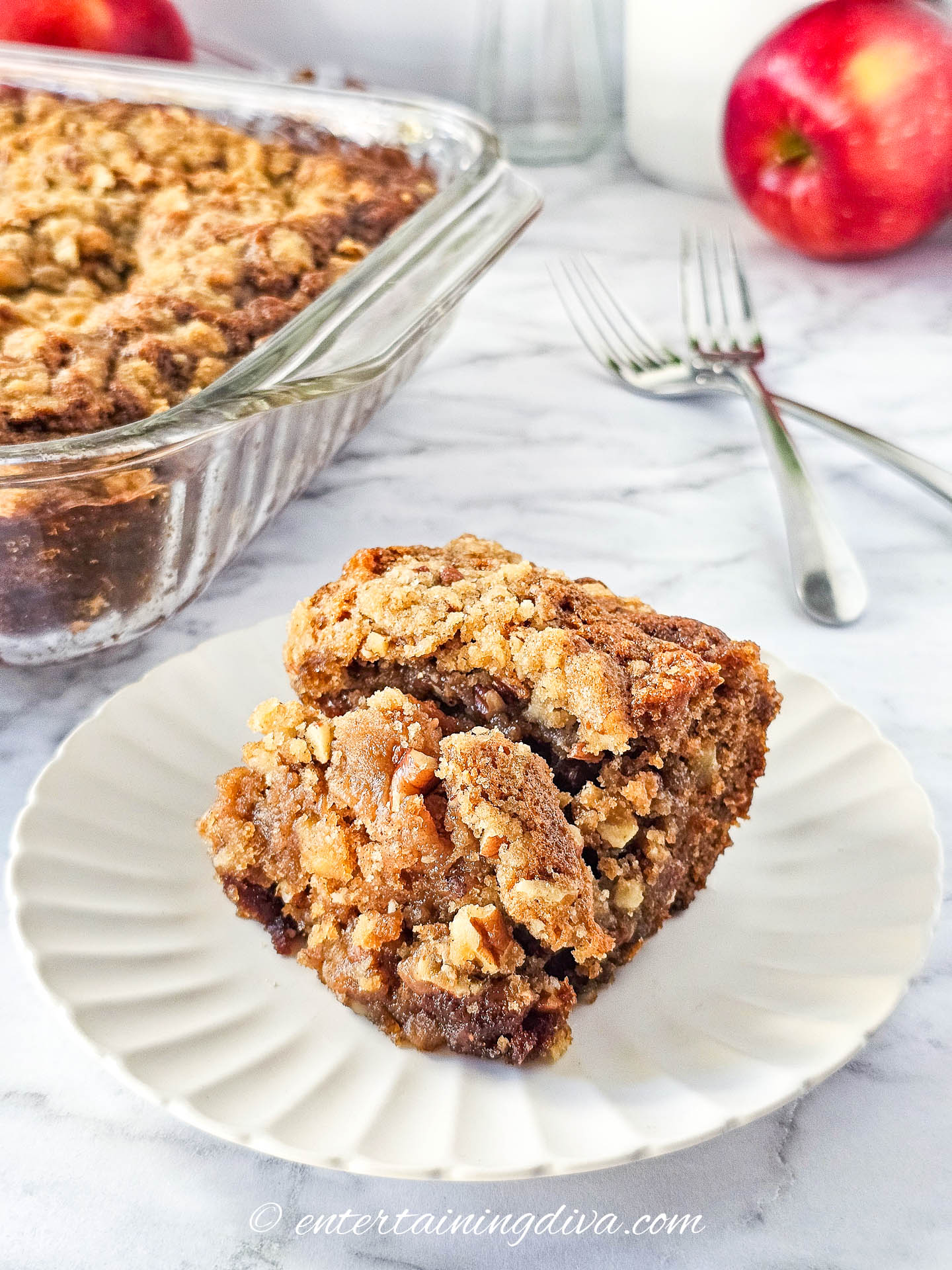 A piece of apple crumb cake on a plate with the rest of the cake in a pan in the background