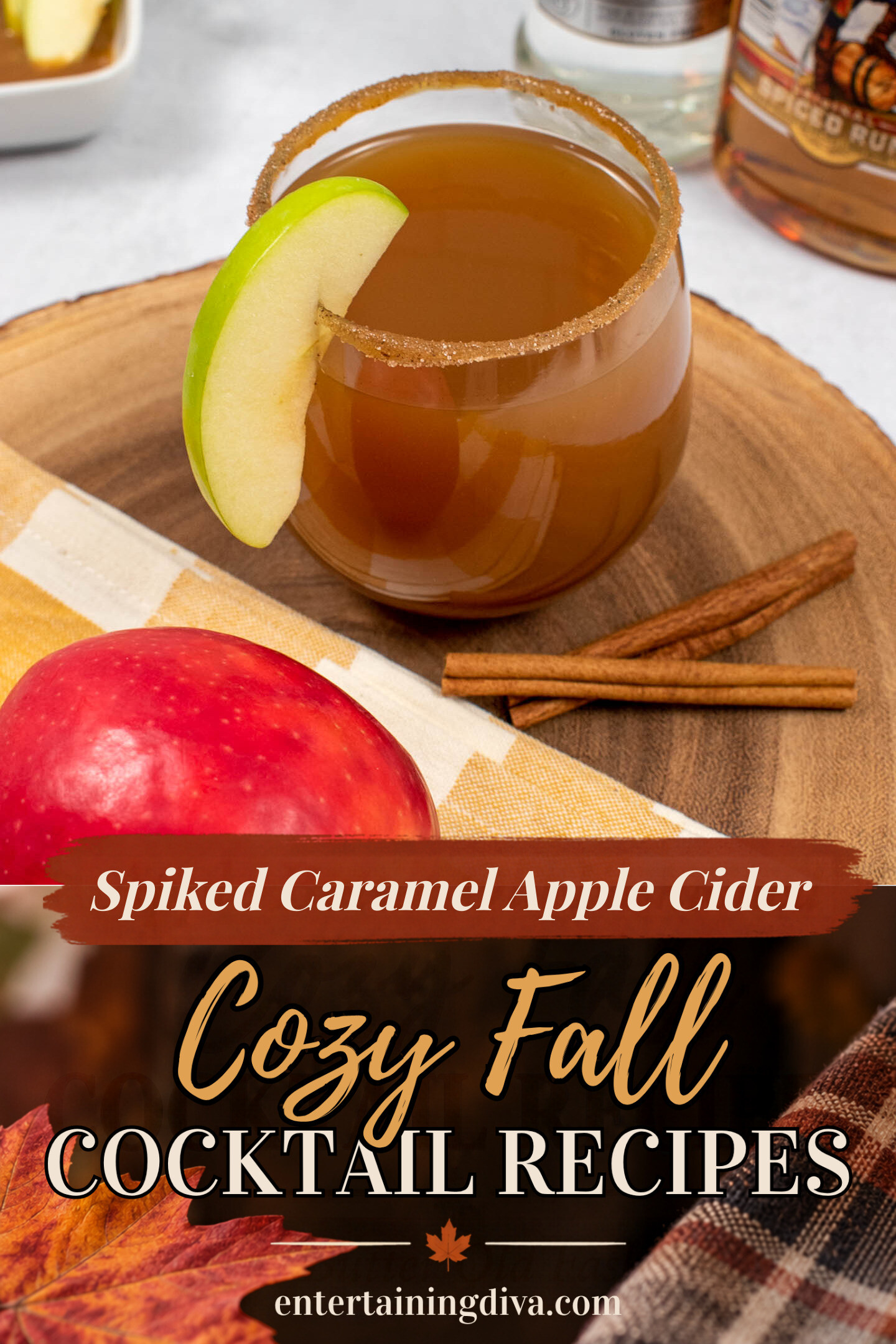 A glass of brown cocktail with a cinnamon-sugar rim and apple slice garnish sits on a wood platter, next to an apple, cinnamon sticks, and rum bottles in the background.