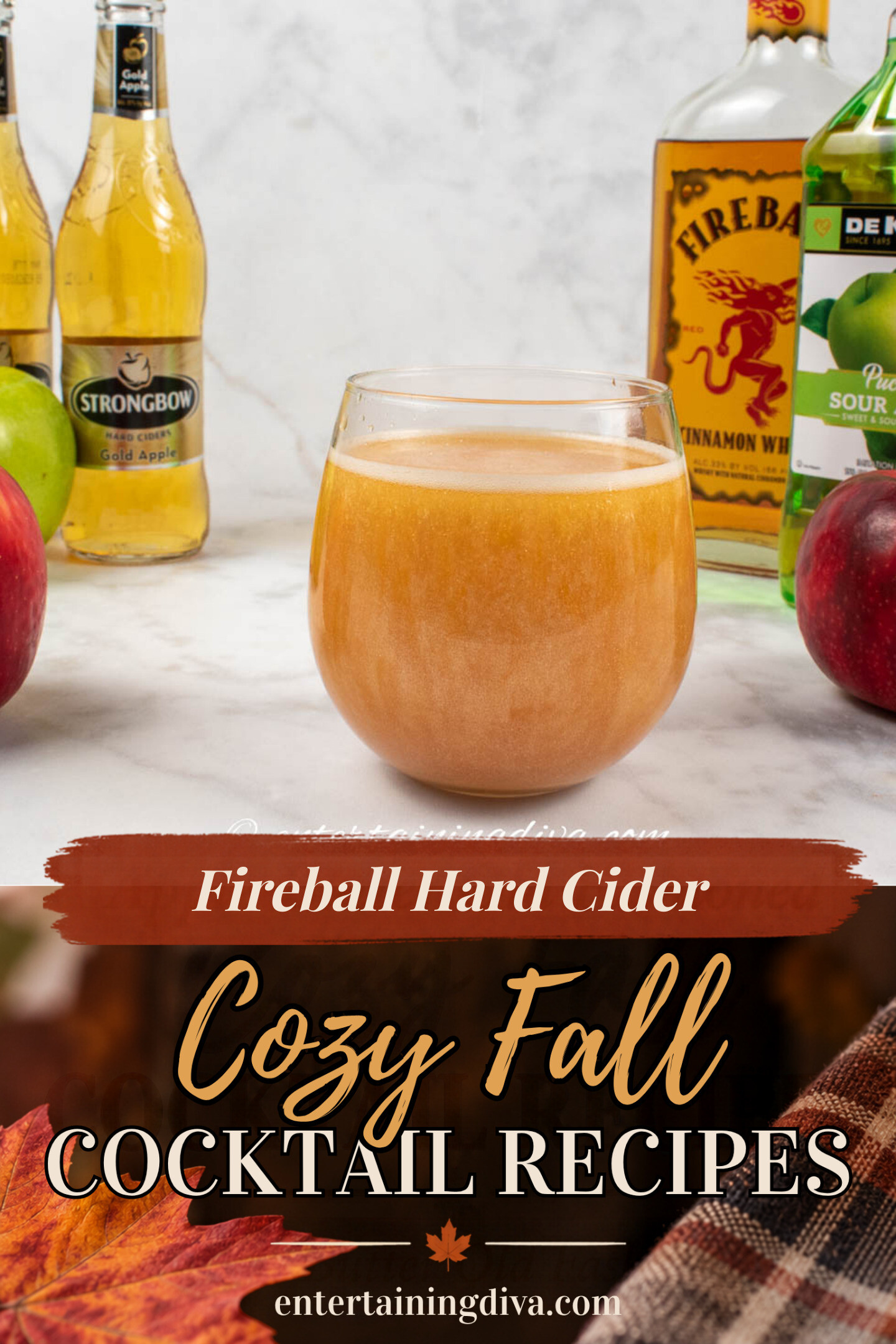 A glass of brownish cocktail is surrounded by two red apples, one green apple, two bottles of hard apple cider, Fireball whiskey, and sour apple liqueur on a white surface.
