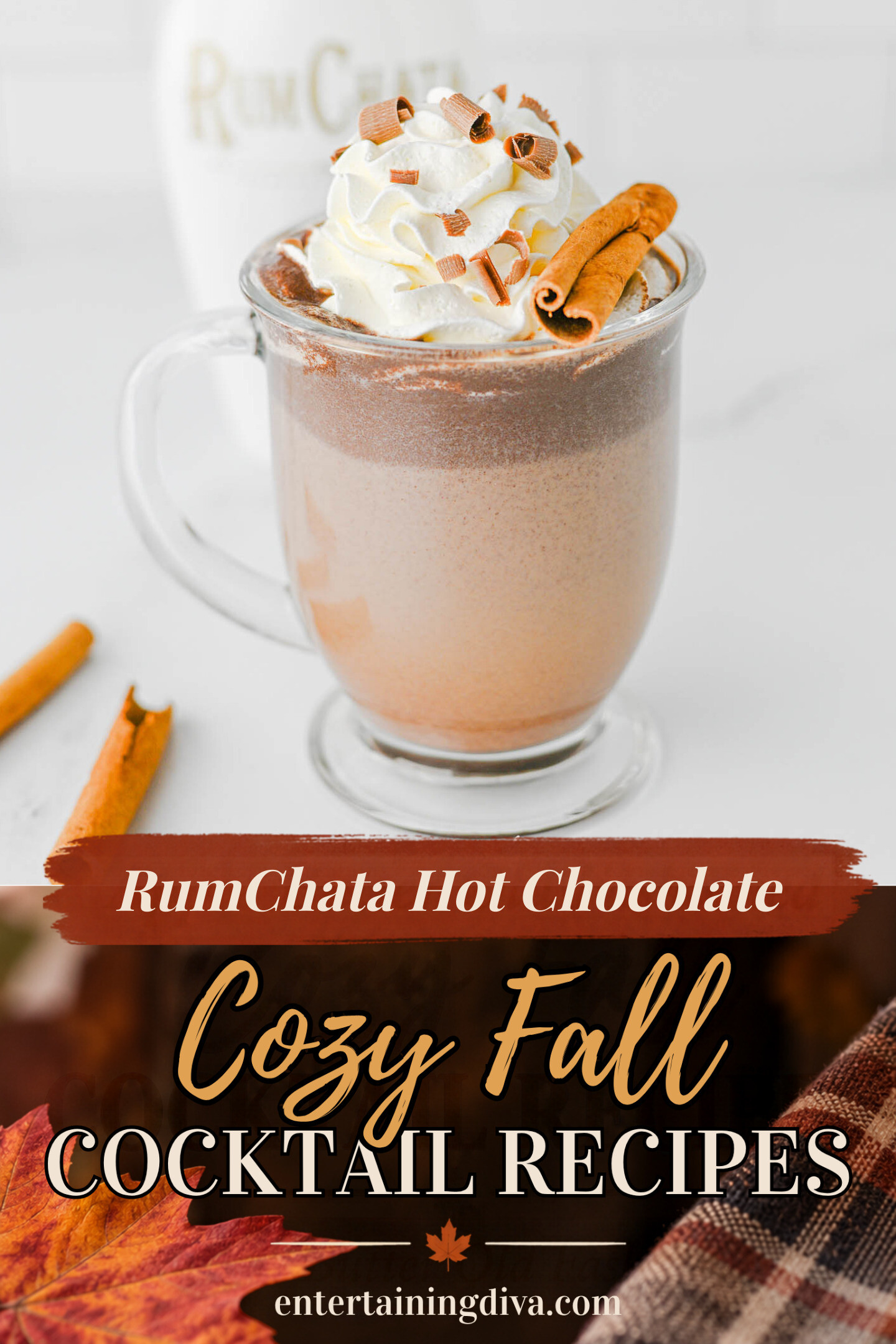 Clear glass mug of hot chocolate topped with whipped cream, chocolate shavings, and a cinnamon stick. A bottle and cinnamon sticks are in the background.