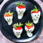 Five strawberries dipped in white chocolate to resemble ghosts, with chocolate dots for eyes and mouths, arranged on a black plate.