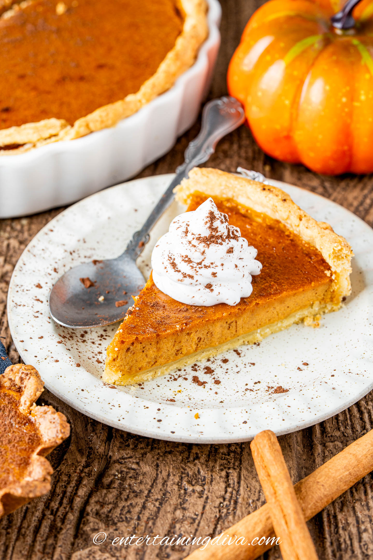 a slice of pumpkin pie on a plate beside cinnamon sticks and a pumpkin