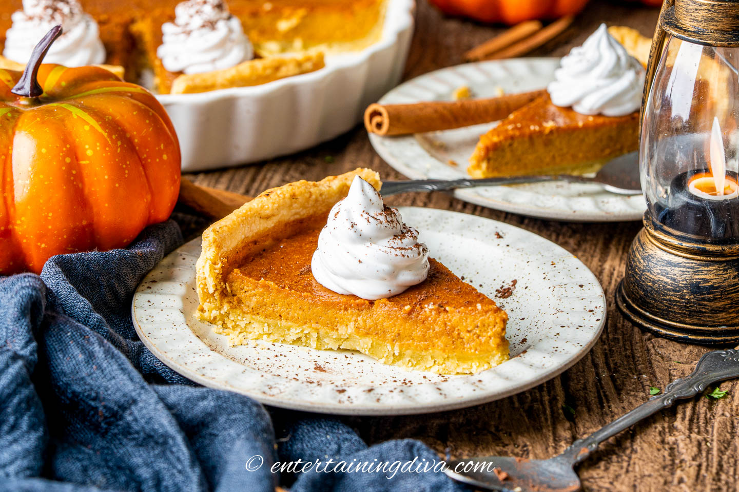 A slice of pumpkin pie on a table with another slice of pie and the rest of the pie in the pan in the background
