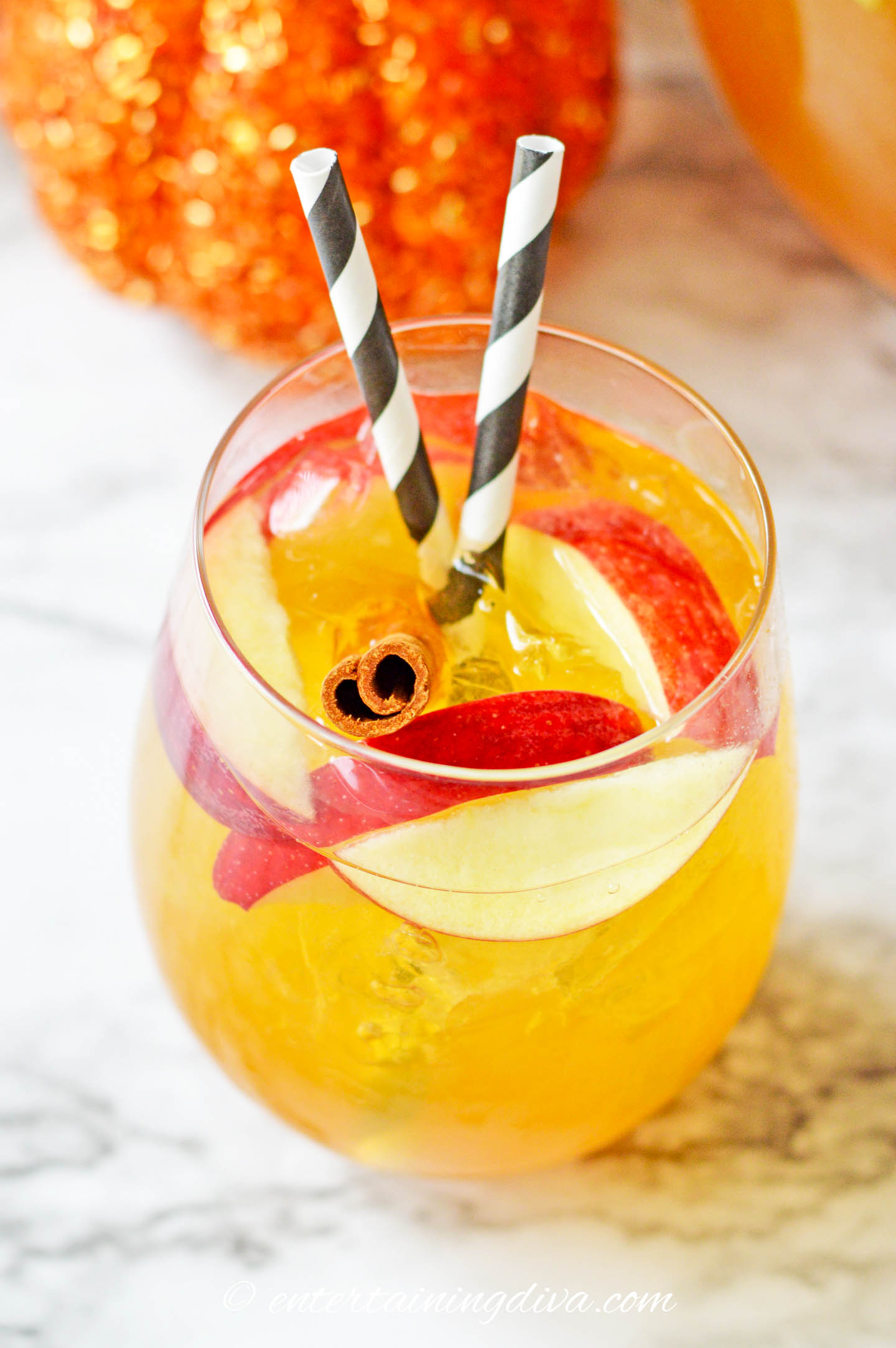 Ice, apple slices, and a cinnamon stick in a glass of pumpkin sangria with two black and white straws