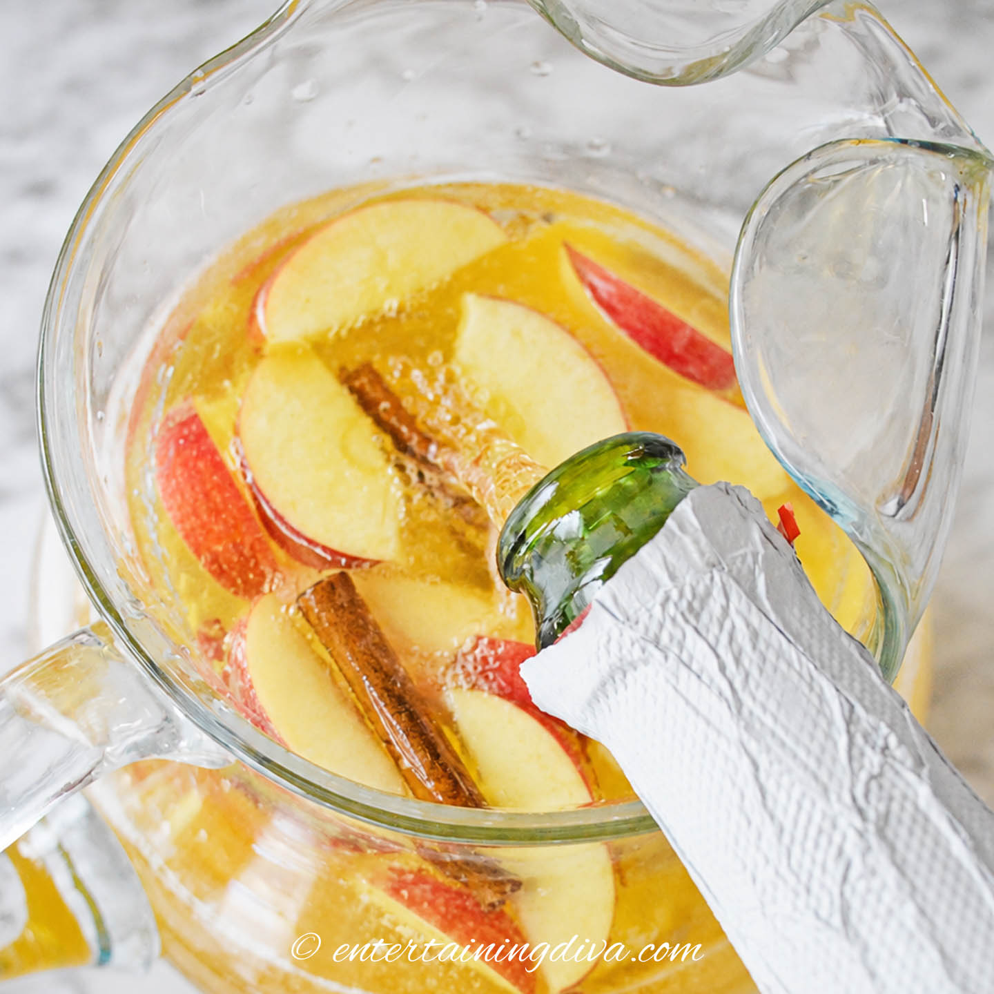sparkling apple juice being poured into a pitcher of pumpkin sangria with apple slices
