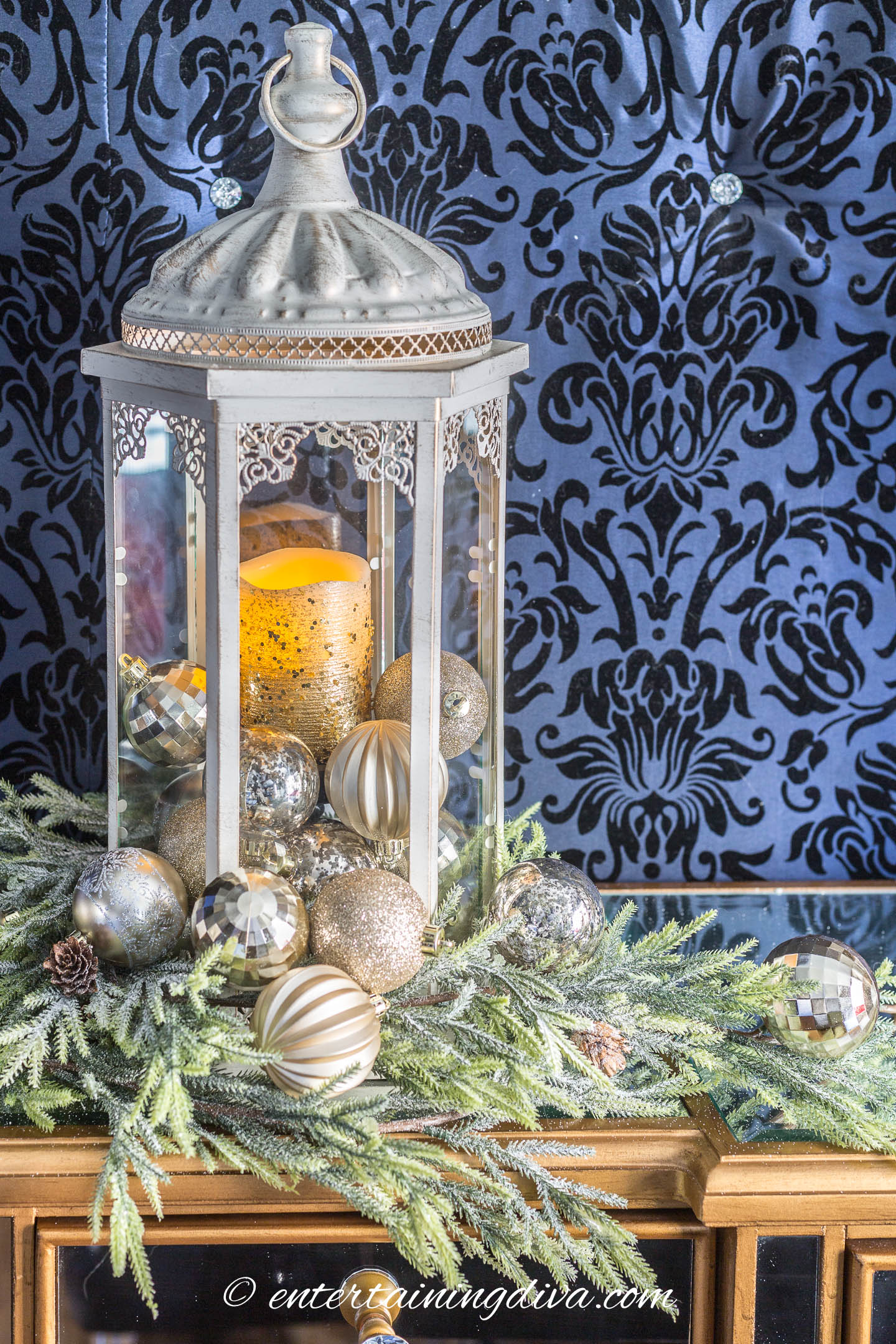 A white lantern filled with ornaments and surrounded by evergreens on a buffet table