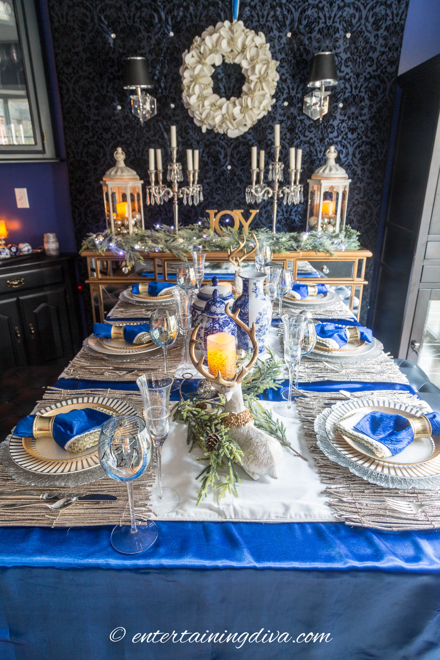 A blue, white and gold Christmas tablescape with a white wreath on the wall behind it