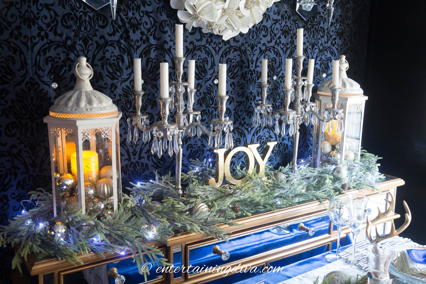 Two white lanterns, two silver candelabras a JOY sign and evergreens on a buffet table