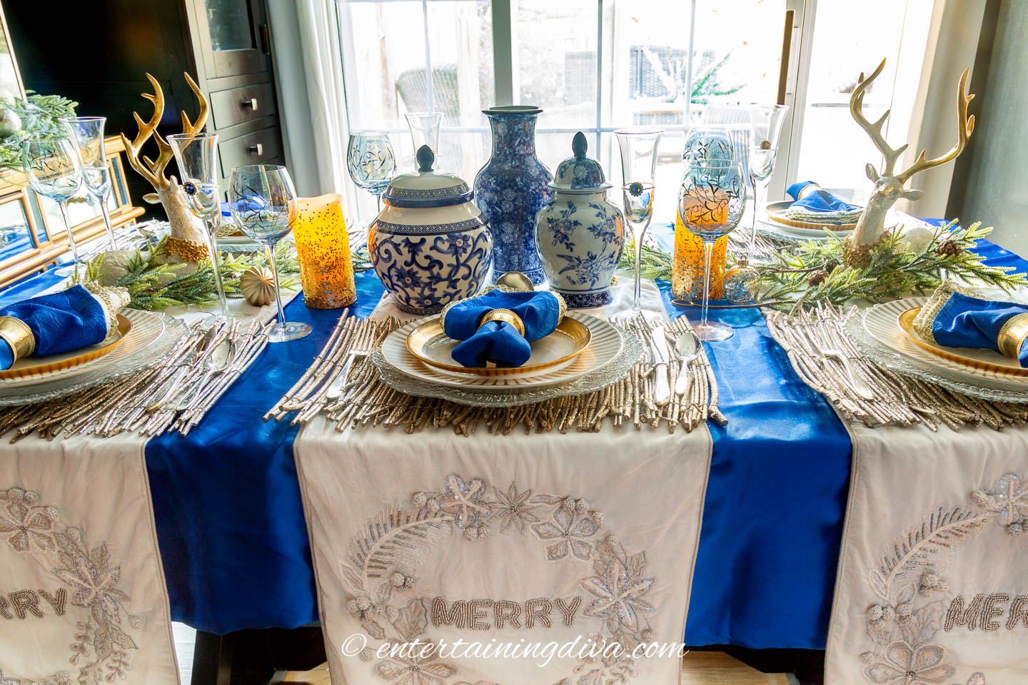 Side view of a blue, white and gold Christmas tablescape with ginger jars, evergreens, candles and deer decorations used as the centerpiece
