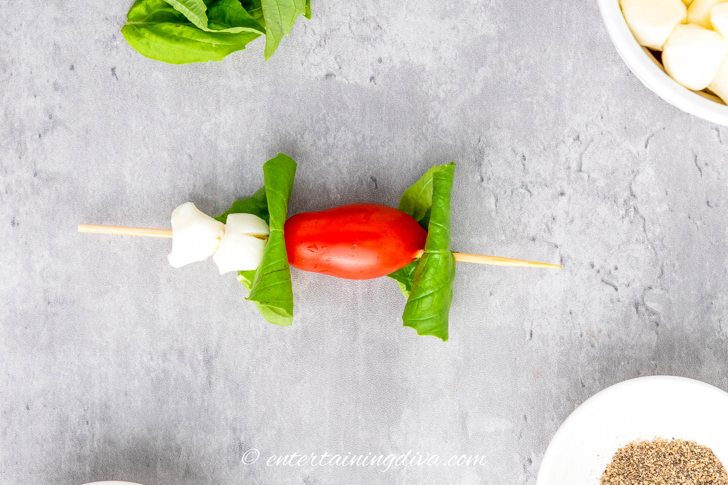 A skewer with 2 mozzarella pearls, 2 folded basil leaves and a grape tomato on a skewer