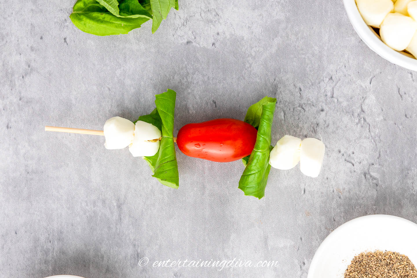 A caprese skewer with 4 small mozzarella pearls, 2 folded basil leaves and a grape tomato on a skewer