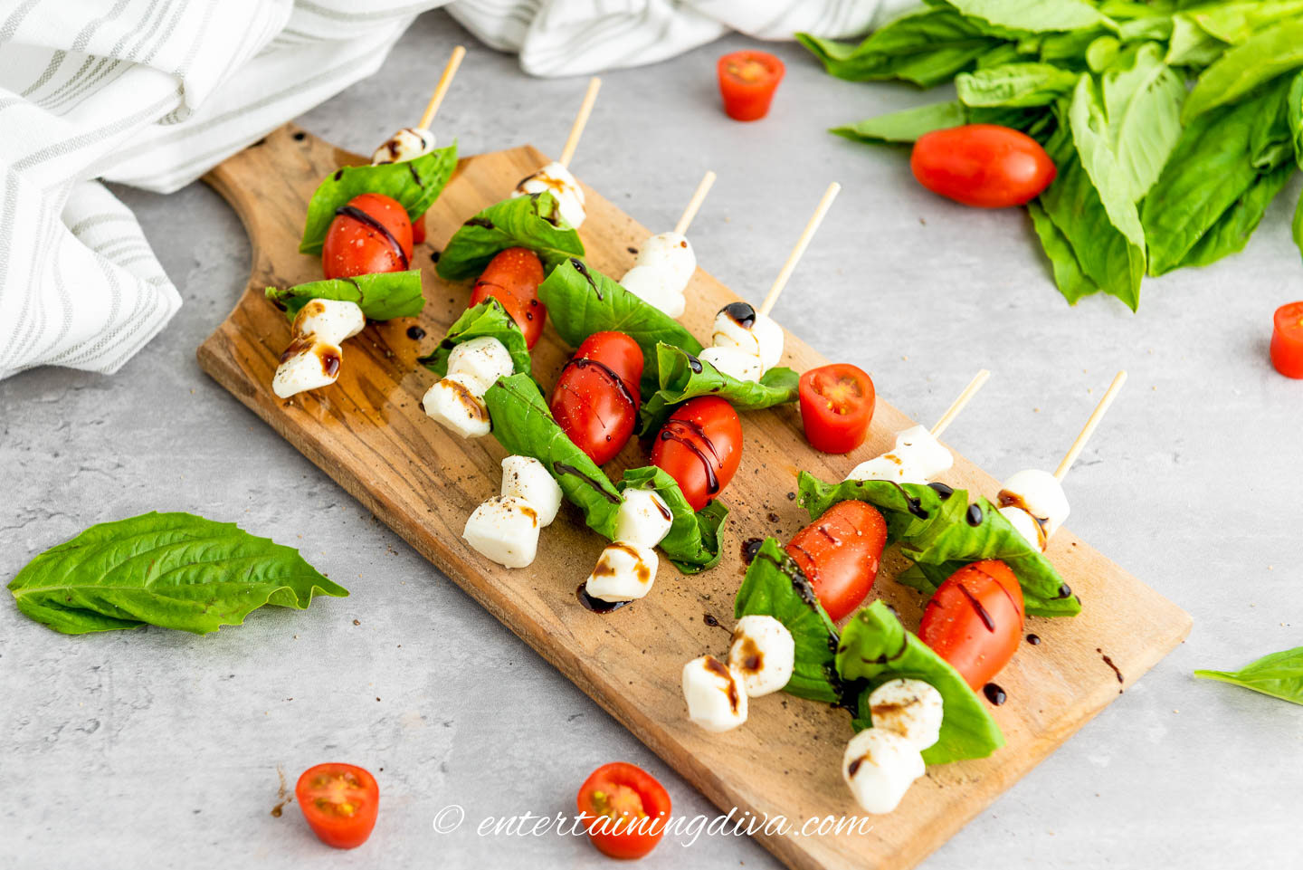 Caprese skewers with balsamic glaze on a serving board with other tomatoes and basil leaves sprinkled around