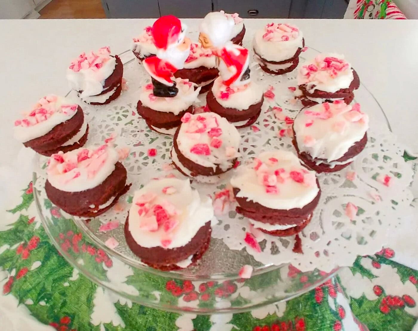 A glass plate holds chocolate sandwich cookies with white frosting and crushed peppermint, topped with two small Santa figurines, on a festive holiday cloth.