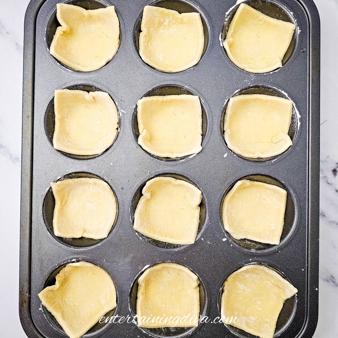 Twelve puff pastry squares pressed into the cavities of a muffin pan.