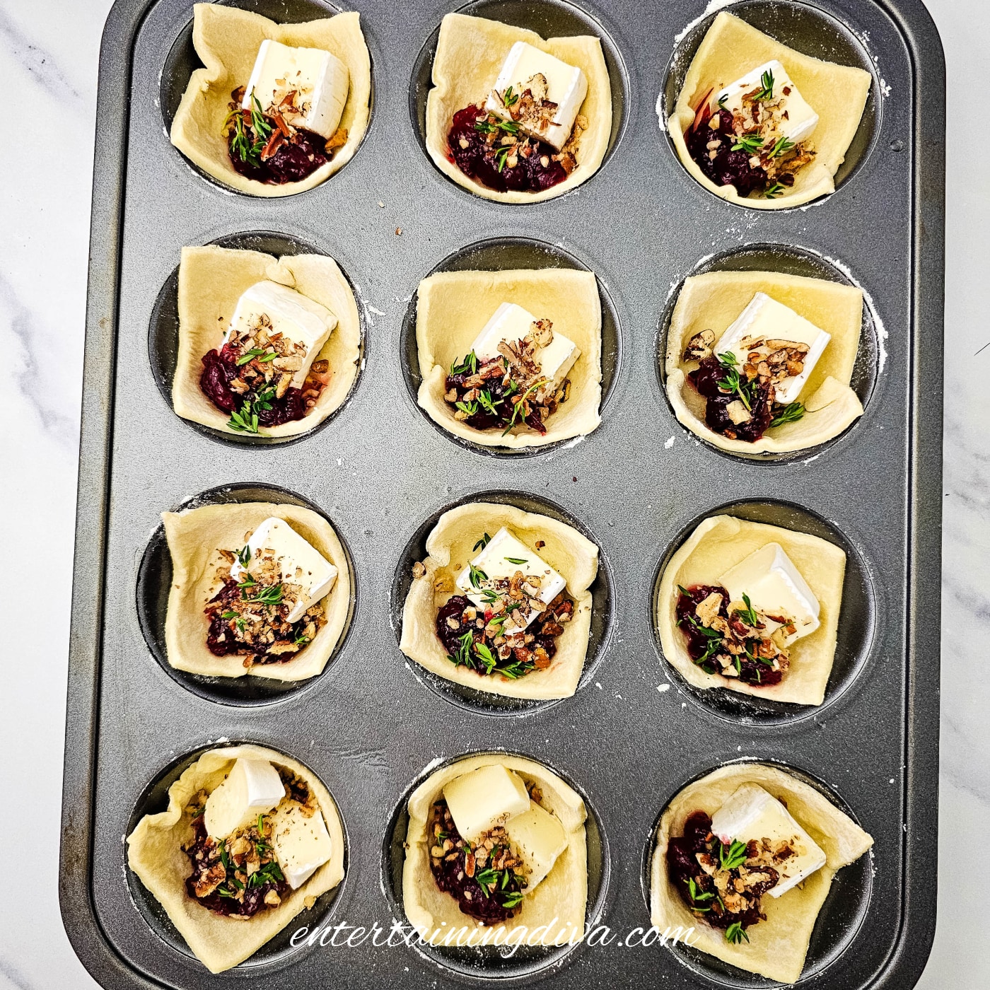 Twelve unbaked cranberry Brie bites sprinkled with chopped pecans and fresh thyme in a muffin pan.
